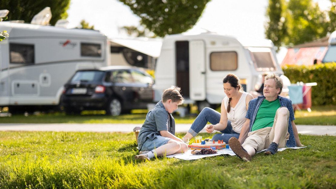 Eine Familie mit Kind sitzt im Gras am Ufer der Fulda entspannt vor ihrem Wohnwagen und spielt gemeinsam.